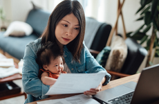 Asian mother holding their child and reviewing paperwork in front of a laptop.