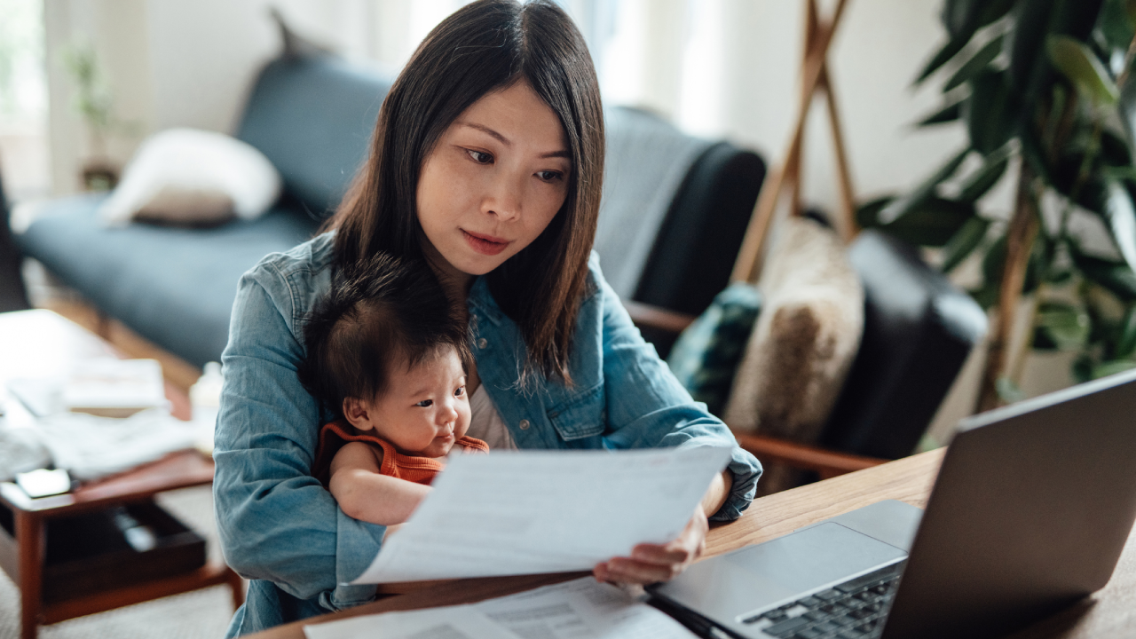 Asian mother holding their child and reviewing paperwork in front of a laptop.