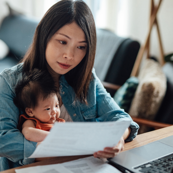 Asian mother holding their child and reviewing paperwork in front of a laptop.