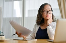 Middle aged Asian business woman holds paperwork and looks at a laptop.