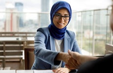 A woman wearing business clothing and a hijab shakes hands across a table.