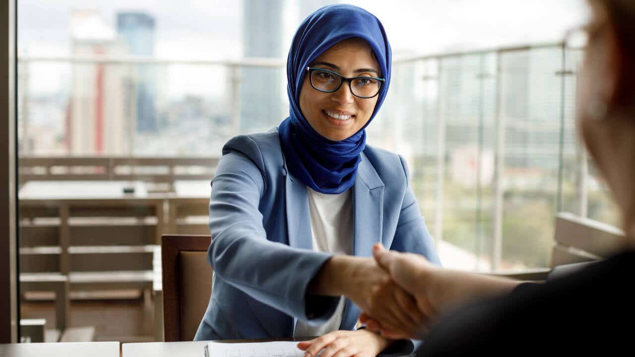 A woman wearing business clothing and a hijab shakes hands across a table.