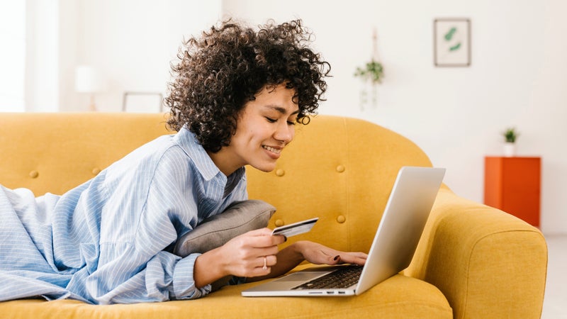 Smiling woman holding credit card using laptop lying on sofa in living room at home