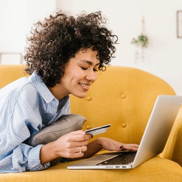 Smiling woman holding credit card using laptop lying on sofa in living room at home