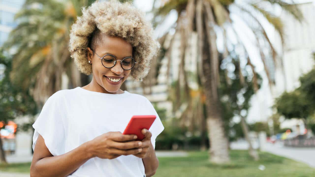 A Black woman with natural hair smiles while using a smartphone outdoors.