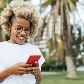 A Black woman with natural hair smiles while using a smartphone outdoors.