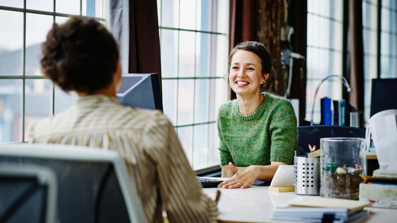 Two female entrepreneurs smile at each other across a desk.