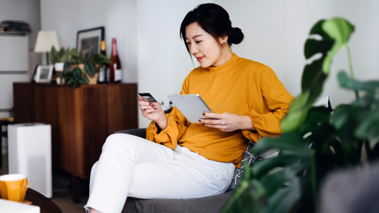 Beautiful smiling young Asian woman sitting on sofa in the living room, managing online banking, handling bank account and financial bills with digital tablet and credit card at home. Convenience and easy banking. Smart banking with technology