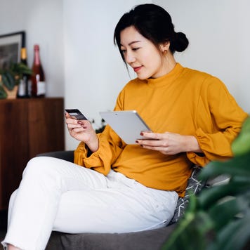 Beautiful smiling young Asian woman sitting on sofa in the living room, managing online banking, handling bank account and financial bills with digital tablet and credit card at home. Convenience and easy banking. Smart banking with technology