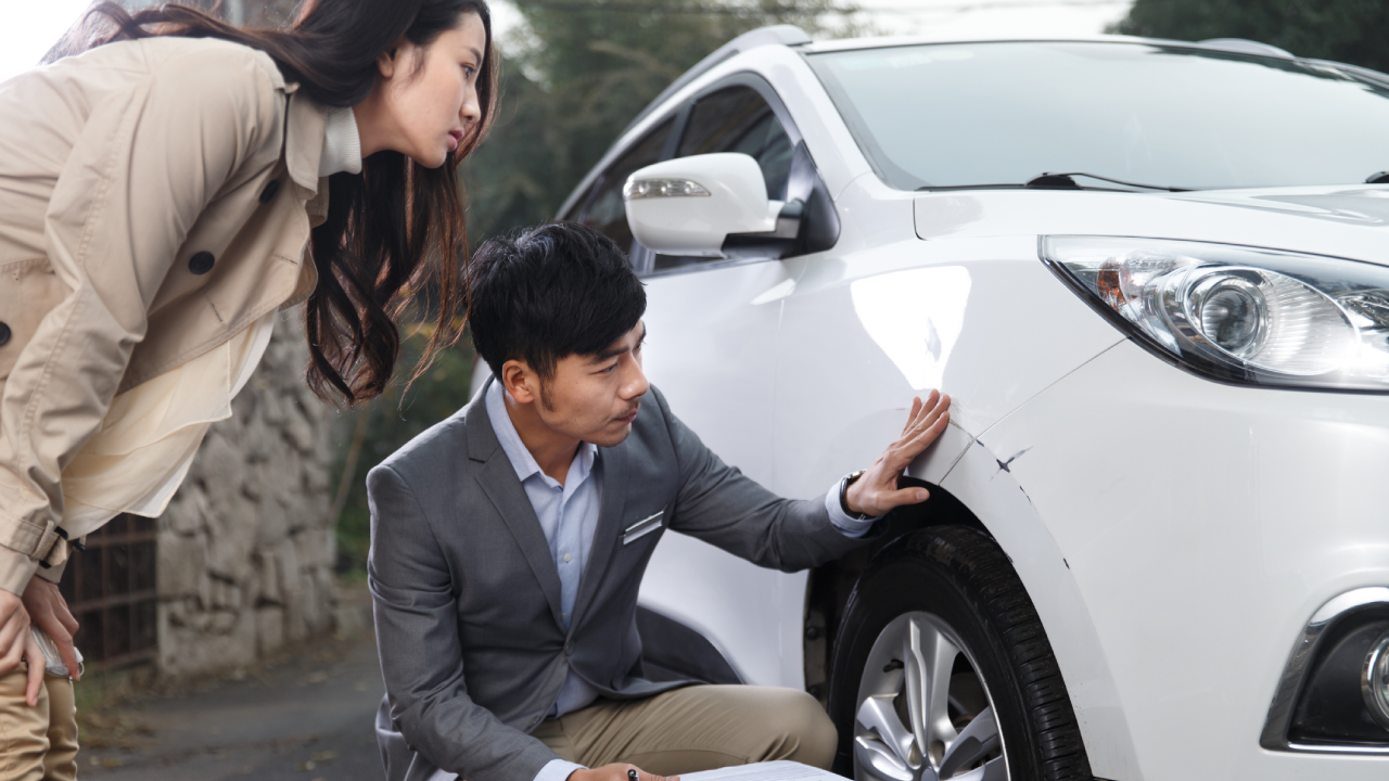 An insurance adjuster assesses the damage to a car while the driver looks on with concern.