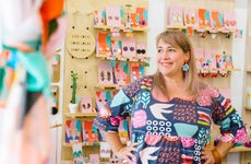 A mature, female business owner smiles in front of a colorful display of jewelry.