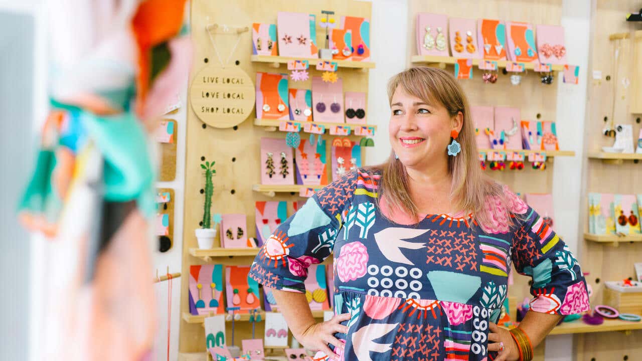 A mature, female business owner smiles in front of a colorful display of jewelry.