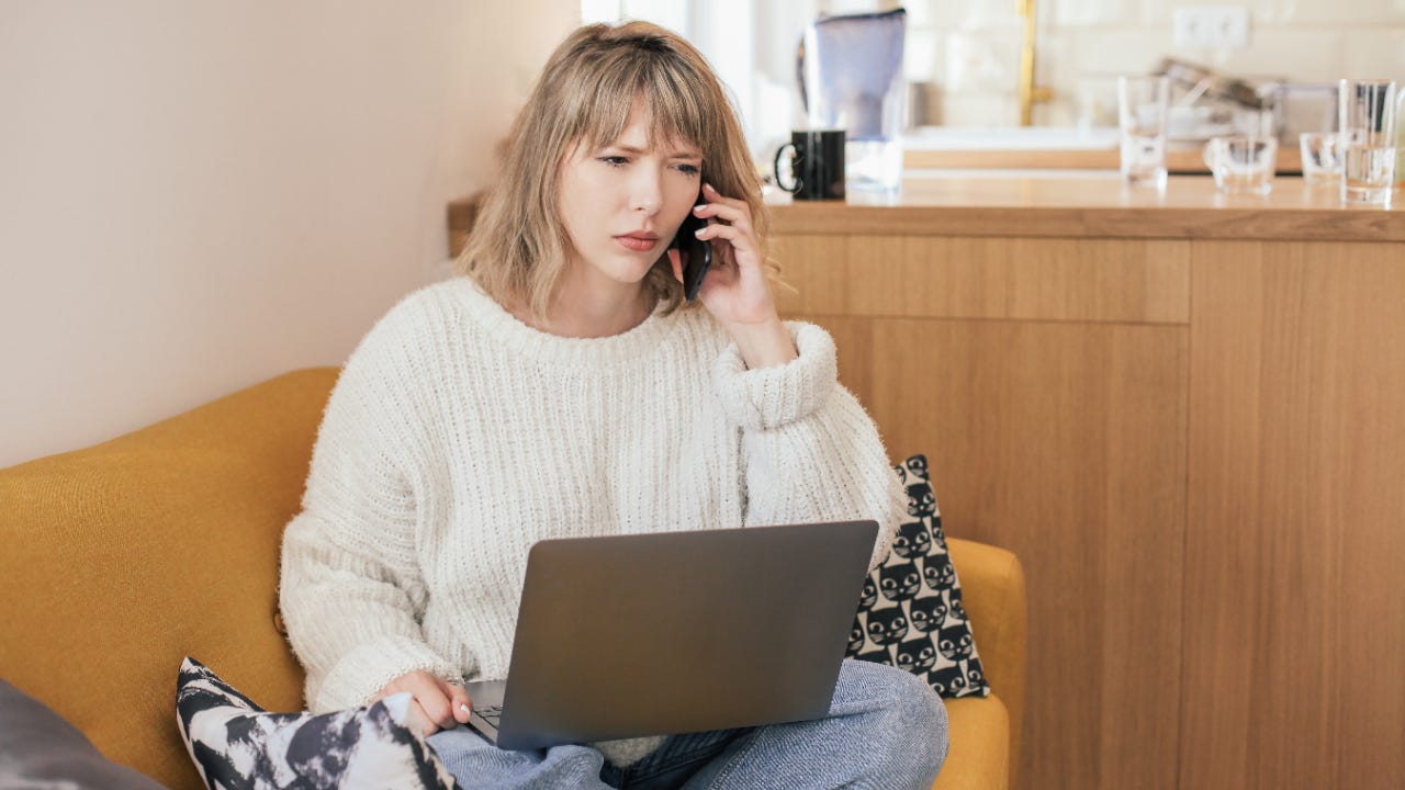 Upset woman on smartphone, sitting on couch with laptop