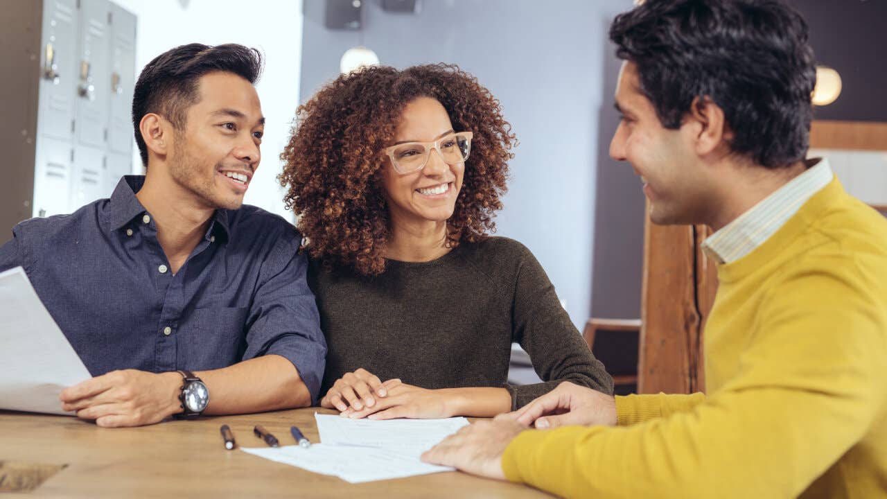 A man and woman smile while speaking to a lender. There's paperwork on the desk between them.