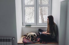 Woman with long hair sitting on window sill using laptop and sleeping pug, looking at snow from the window