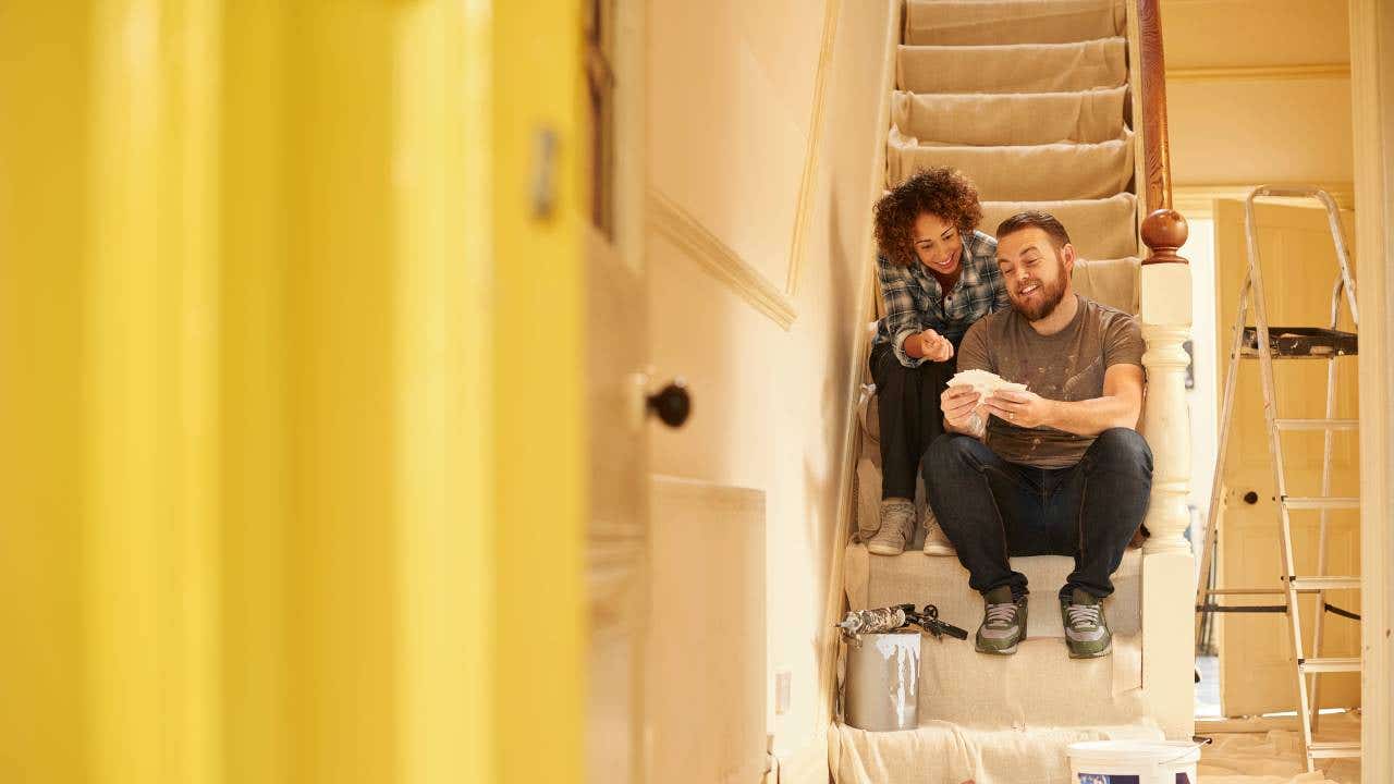 A young couple doing up their hallway in the old house they have bought . They are sitting on the dust sheets on the stairs and looking through the paint sample cards choosing the final color for the beige hall.