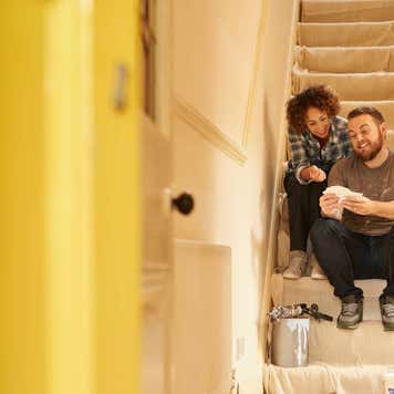 A young couple doing up their hallway in the old house they have bought . They are sitting on the dust sheets on the stairs and looking through the paint sample cards choosing the final color for the beige hall.