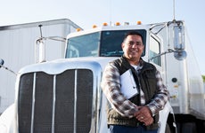 A semi-truck driver stands in front of his truck, smiling