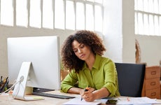 A Black, female business owner with natural hair sits at a computer desk, highlighting documents.