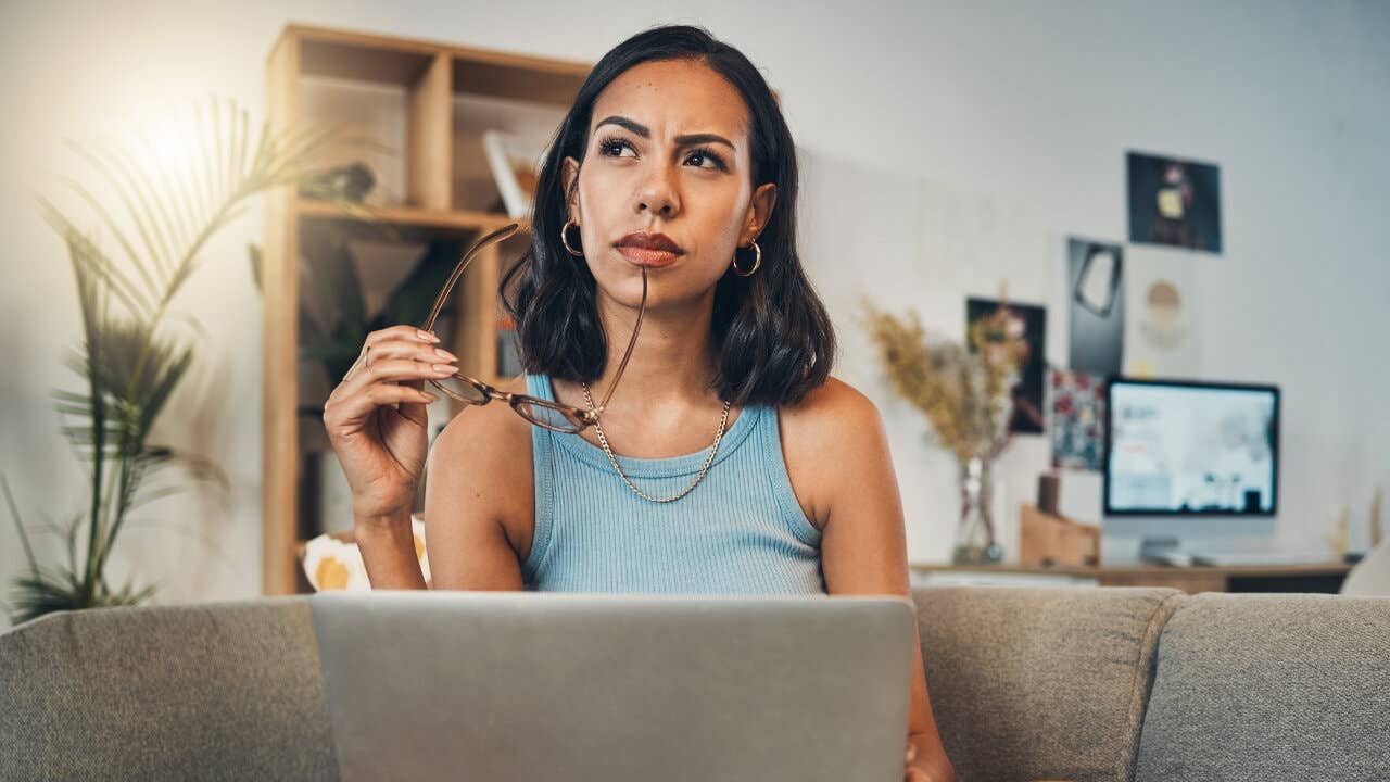 A woman sitting in front of a laptop looks pensively into the distance.