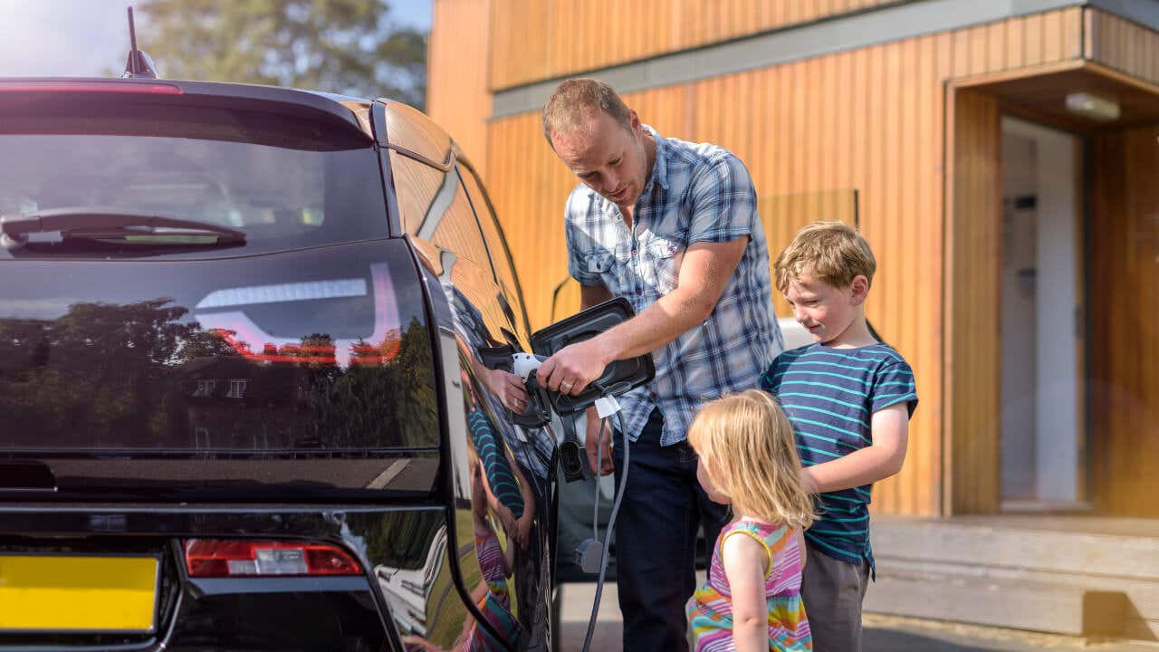 A father plugs in his electric vehicle at home while his two small children watch.