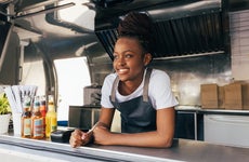 A Black woman with braided hair smiles while leaning against the counter of a food truck.
