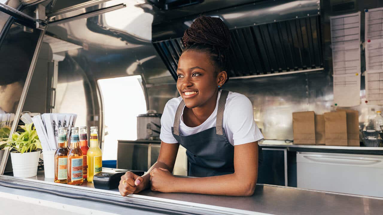 A Black woman with braided hair smiles while leaning against the counter of a food truck.