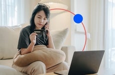 design image of a woman on the phone holding a credit card in one hand and sitting in front of a laptop on a couch