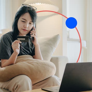 design image of a woman on the phone holding a credit card in one hand and sitting in front of a laptop on a couch