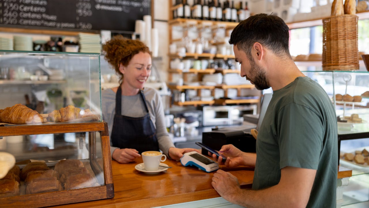 A female business owner serves a cup of coffee over to a male customer who uses his phone to pay.