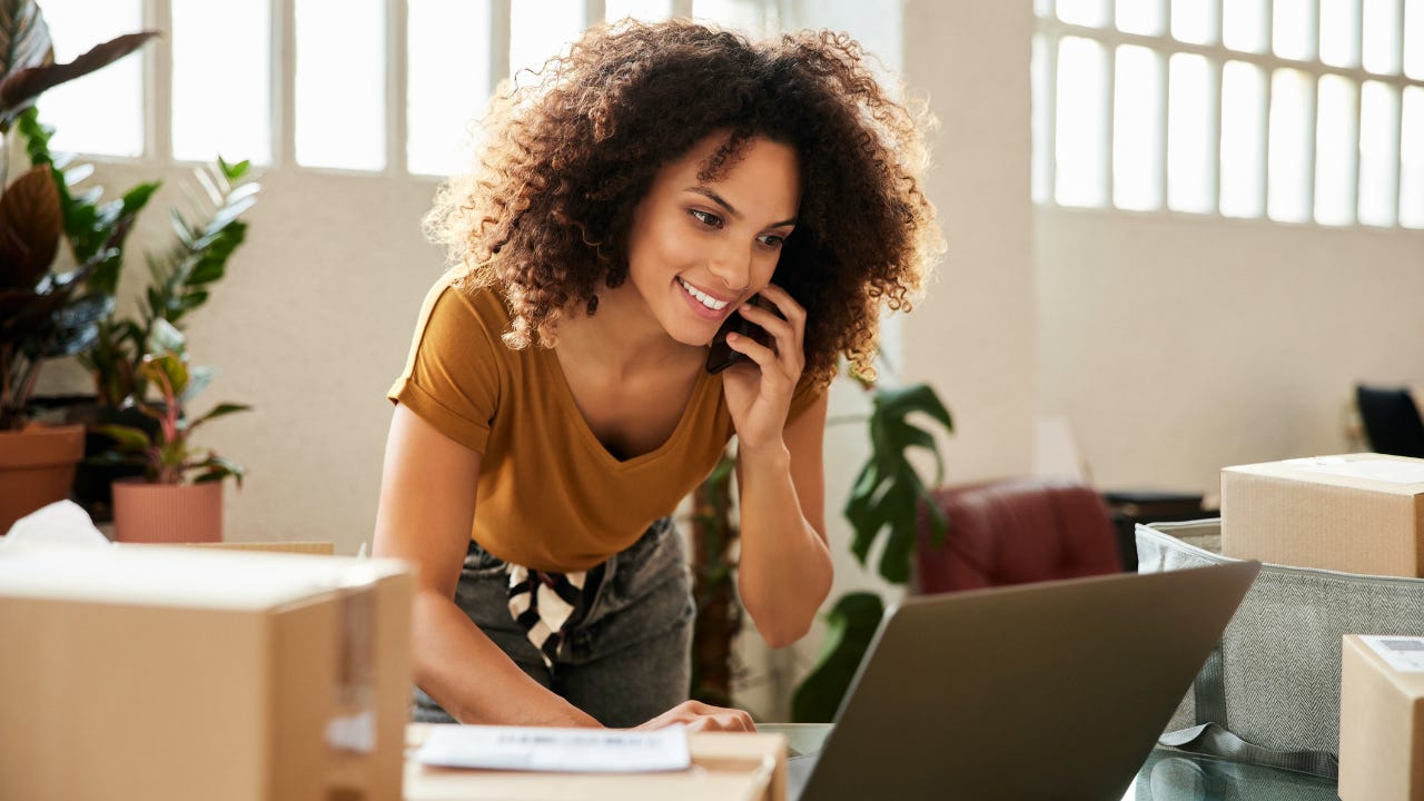 A woman leans over a desk in her office, talking on the phone and looking at her laptop.