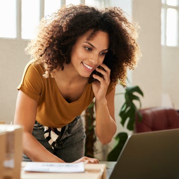 A woman leans over a desk in her office, talking on the phone and looking at her laptop.