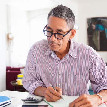 A man with gray hair sits at a desk, using a calculator and a pen and paper.