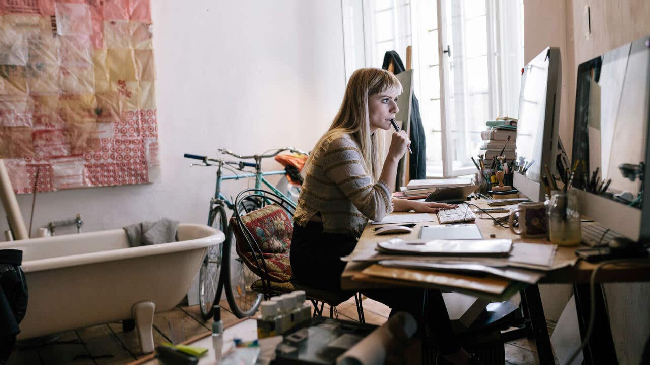 A woman sitting at a cluttered desk in a small apartment looks at a computer screen.