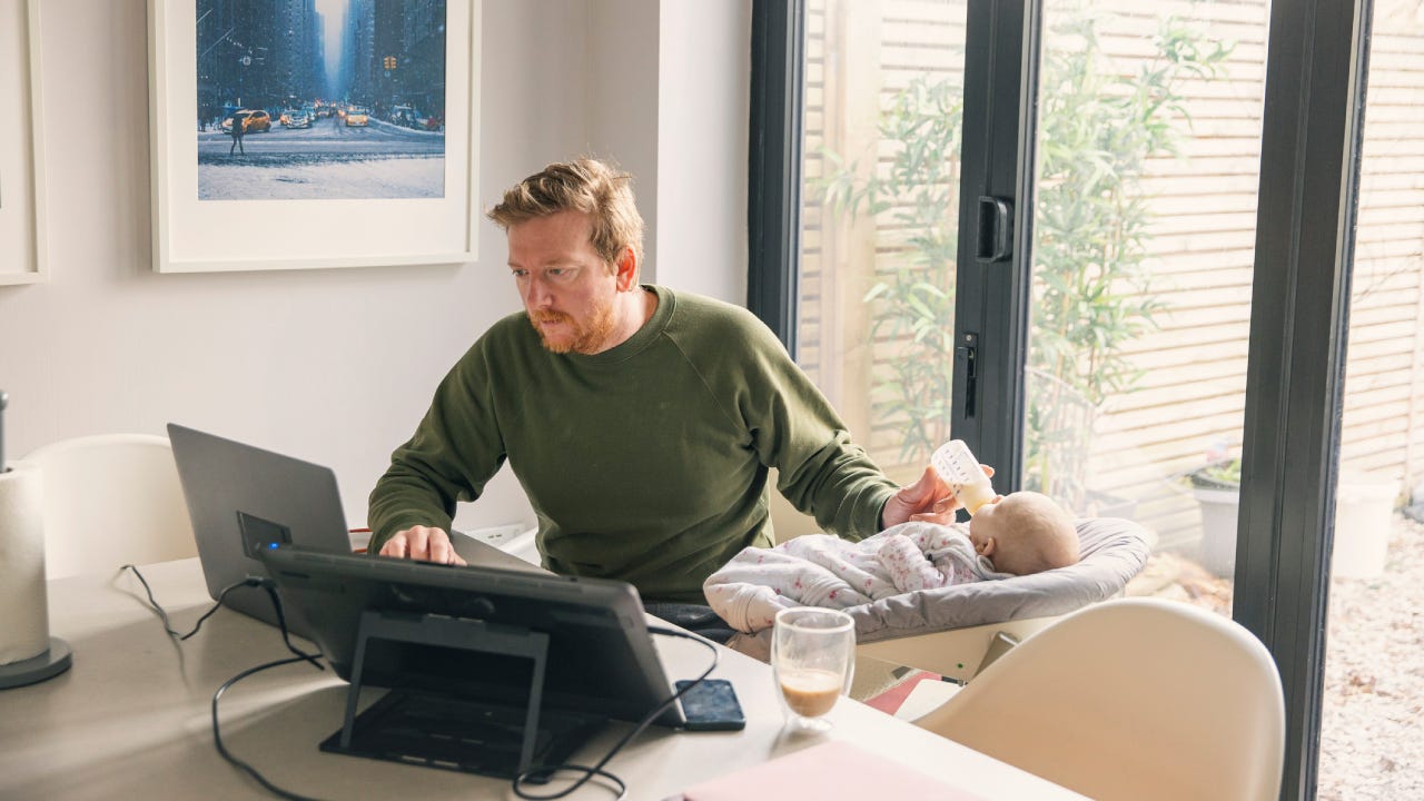 Man working from home dining table whilst feeding his baby daughter