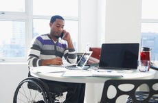 A Black business man sits at a table talking on a phone and looking at paperwork. He is sitting in a wheelchair.