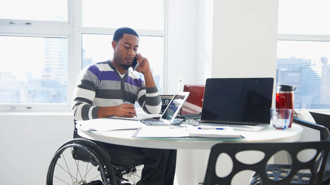 A Black business man sits at a table talking on a phone and looking at paperwork. He is sitting in a wheelchair.