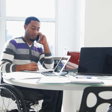 A Black business man sits at a table talking on a phone and looking at paperwork. He is sitting in a wheelchair.