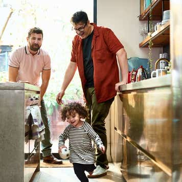 Two fathers watching their toddler playing with ball in kitchen