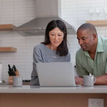 A mixed race couple use a laptop computer to pay bills online, manage budget and prepare tax documents. They are in the kitchen of their home.