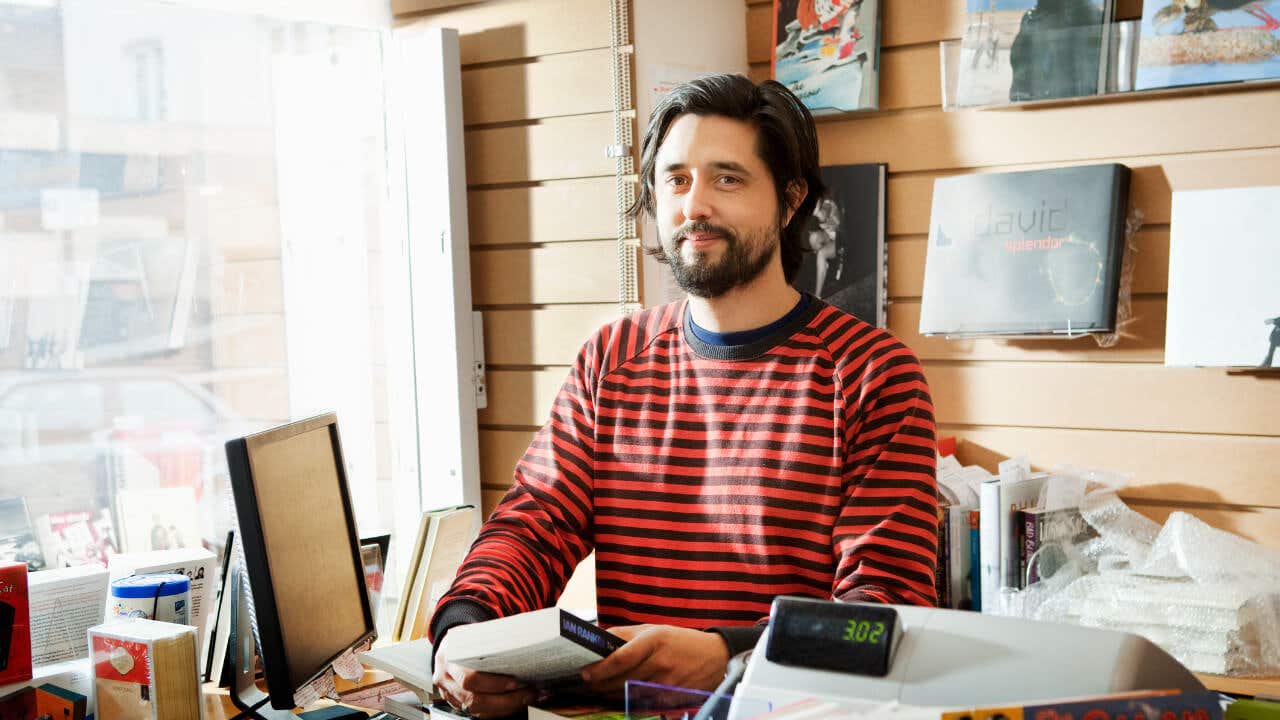 A bookshop owner stands behind his counter.