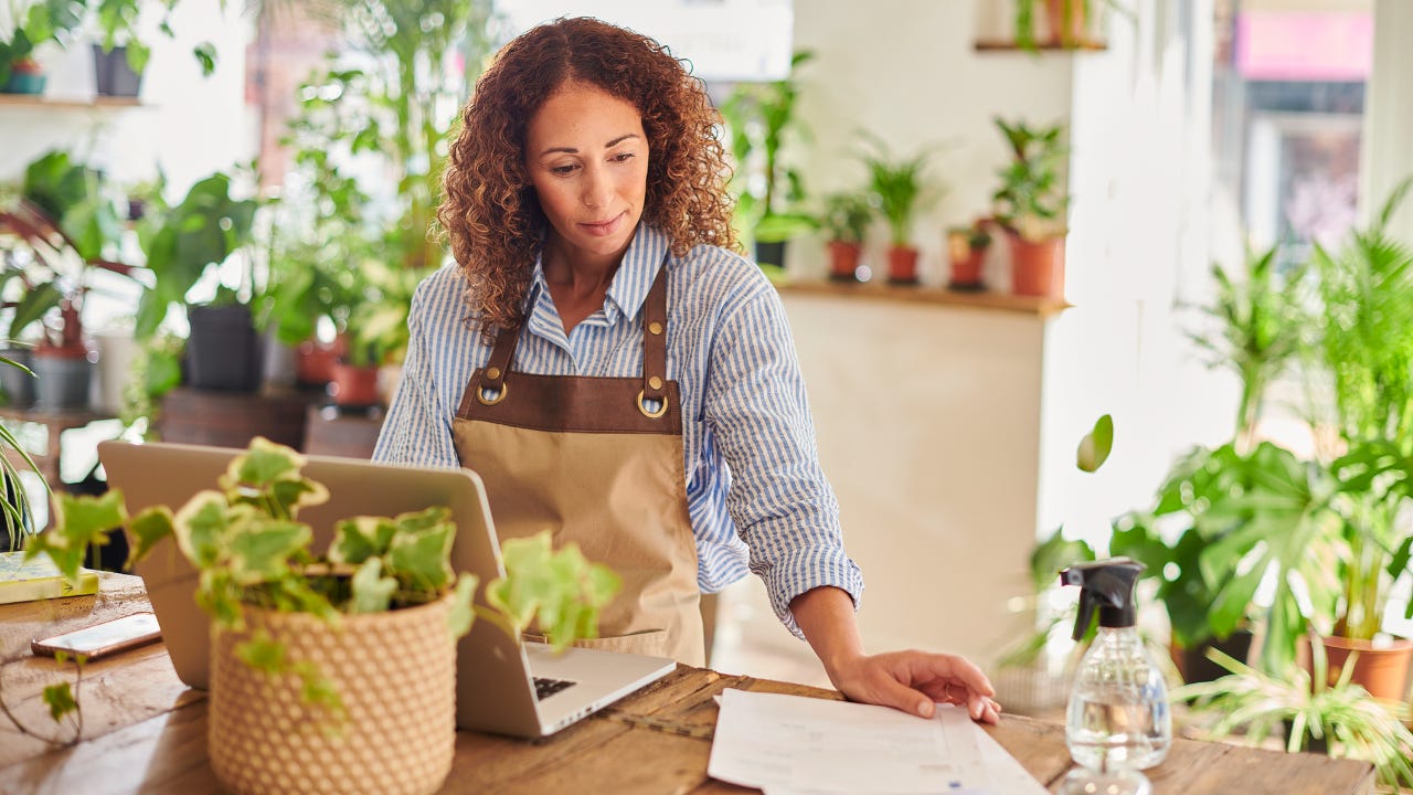 A small business owner stands in her flower shop, looking over her business accounts on her laptop.