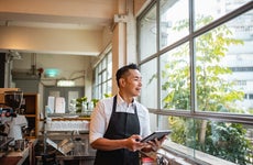 A business owner holding a tablet and smiling as he looks out the window of his cafe.