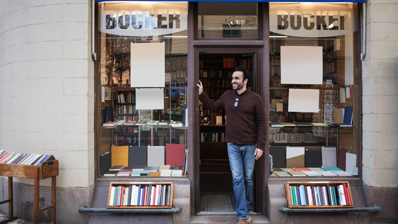 A bookstore owner stands outside his store, smiling and looking down the road.