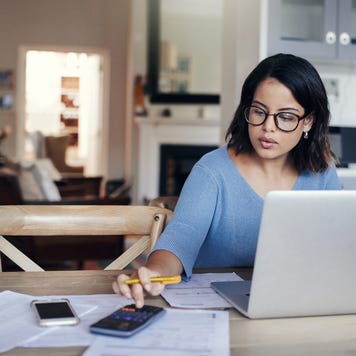 A woman sits at her kitchen table with her laptop, spreadsheets and calculator