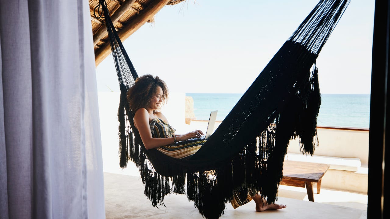 Wide shot of woman working on laptop while relaxing in hammock on deck of luxury tropical villa overlooking ocean