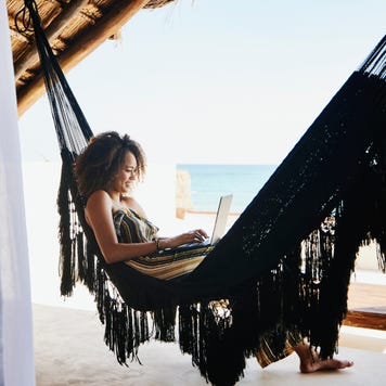 Wide shot of woman working on laptop while relaxing in hammock on deck of luxury tropical villa overlooking ocean