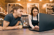 Two young business owners lean over a counter in their coffee shop, looking at a laptop.