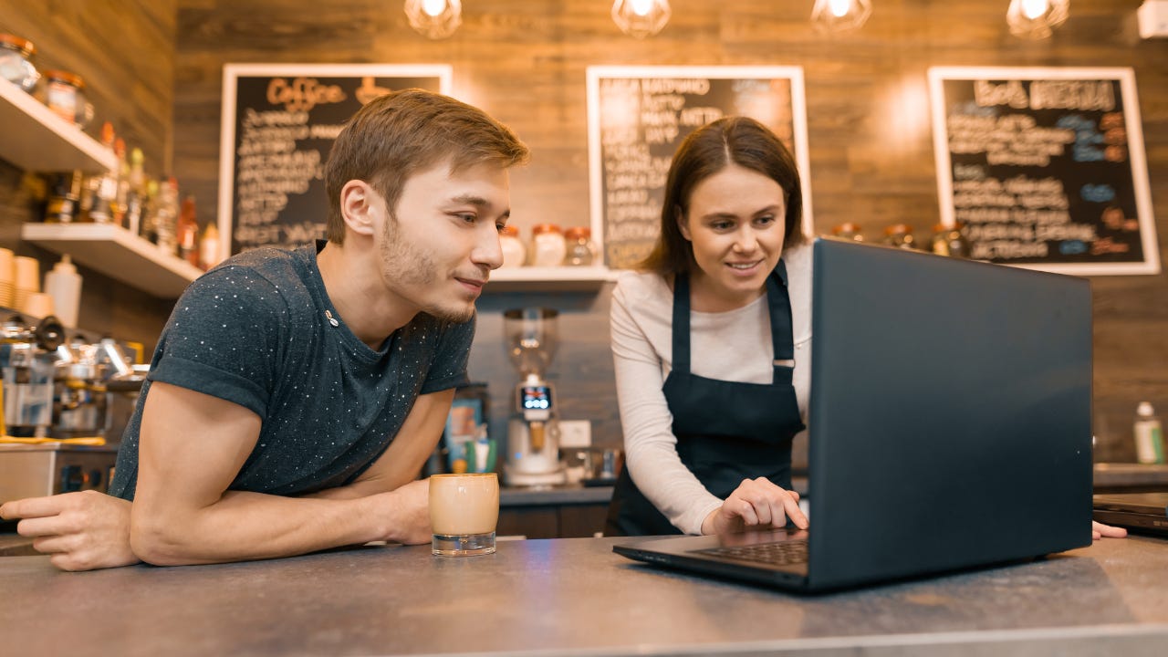 Two young business owners lean over a counter in their coffee shop, looking at a laptop.