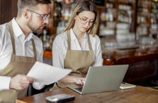 Two cafe owners wearing aprons stand at a table and look over business reports.